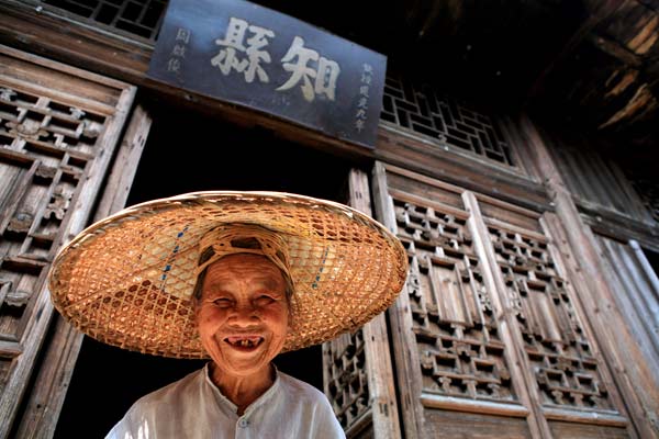 A woman in her 80s poses in front of her house. The wooden board hung above the gate indicates her ancestor used to work as the county head. Huo Yan / China Daily